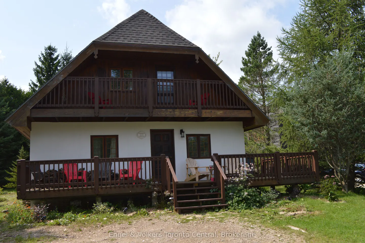 Two-story house with white siding, brown trim, and a brown shingled roof. The house has two decks with red chairs. Trees surround the house.