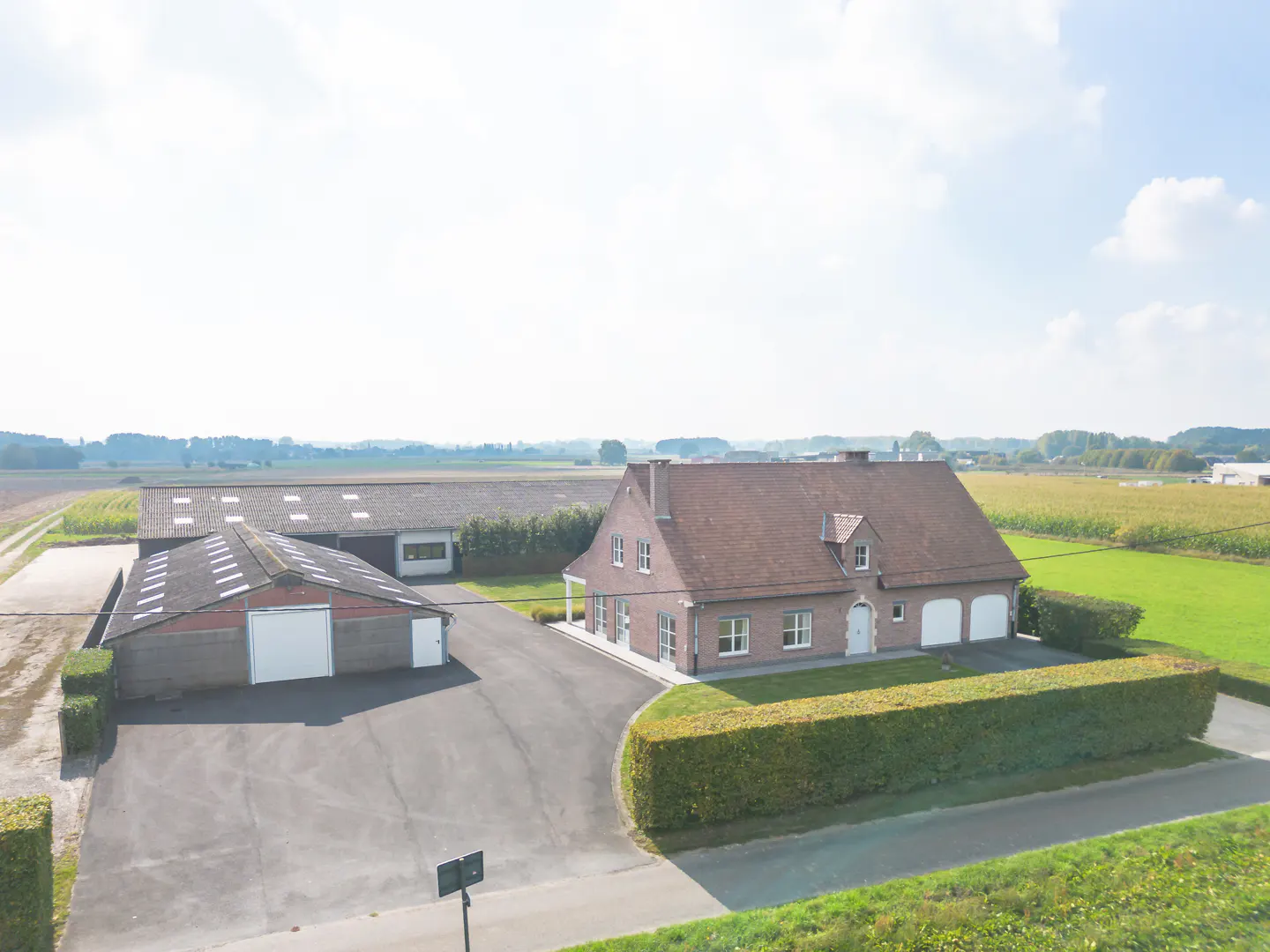 Aerial view of a brick house with a red roof, a barn, and a long driveway surrounded by green fields.