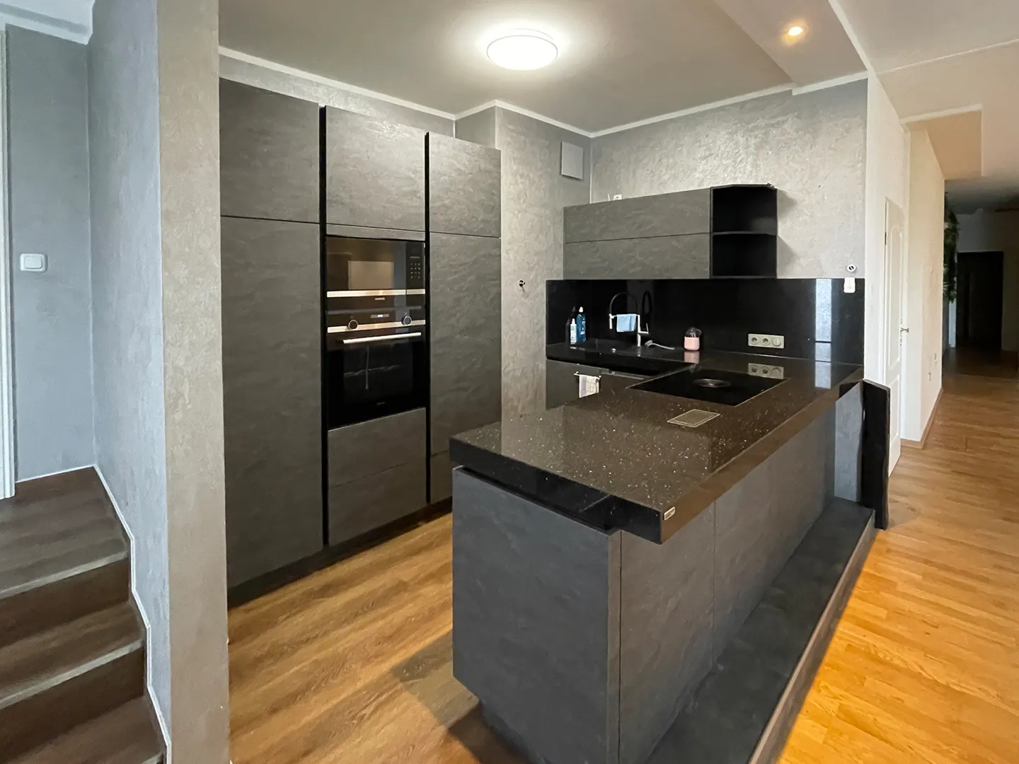 Modern kitchen with gray cabinets, black countertops, and wood floors. A staircase is visible on the left.