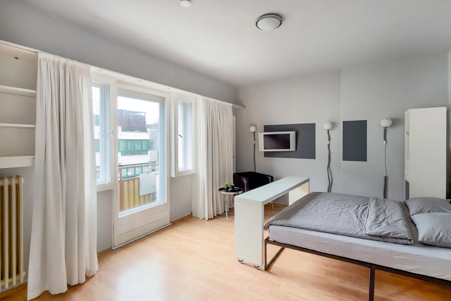 Bright studio apartment with a bed, TV, and large windows with white curtains. The walls are gray and the floor is light wood.