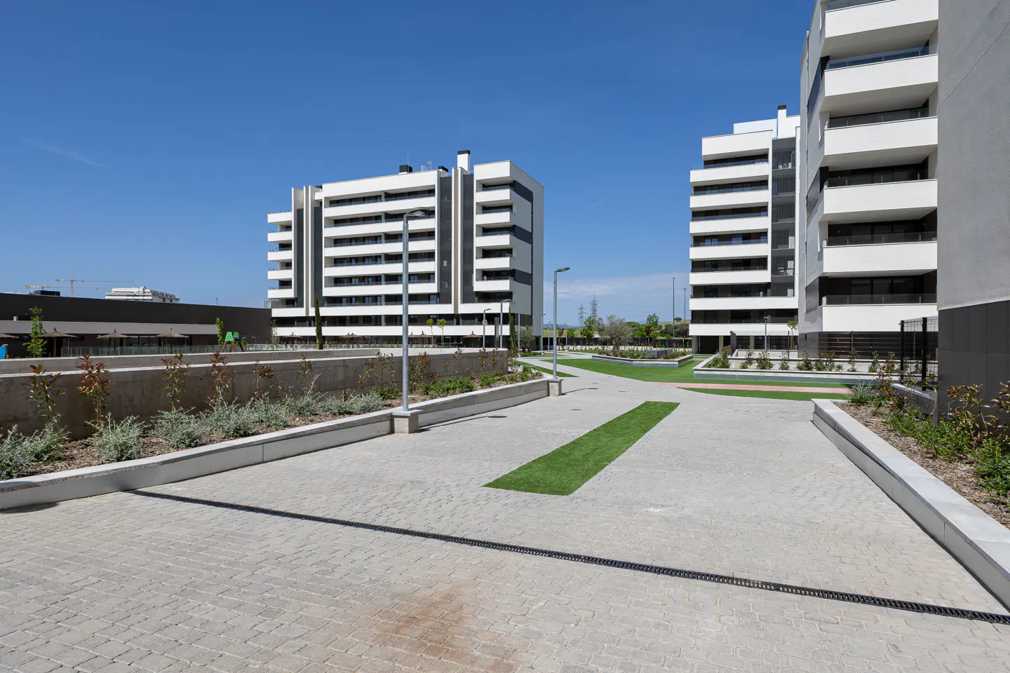Modern white apartment buildings with balconies, viewed from a paved courtyard with grass accents under a clear blue sky.