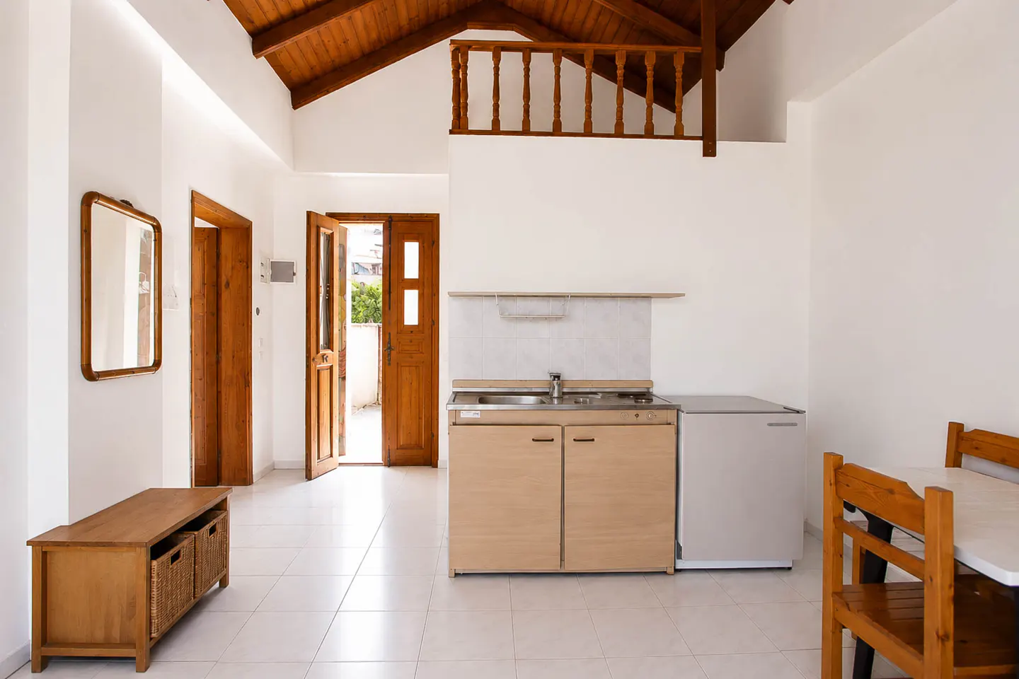 Studio apartment with white walls, tiled floors, a small kitchenette, wooden loft railing, and an open door to a balcony.