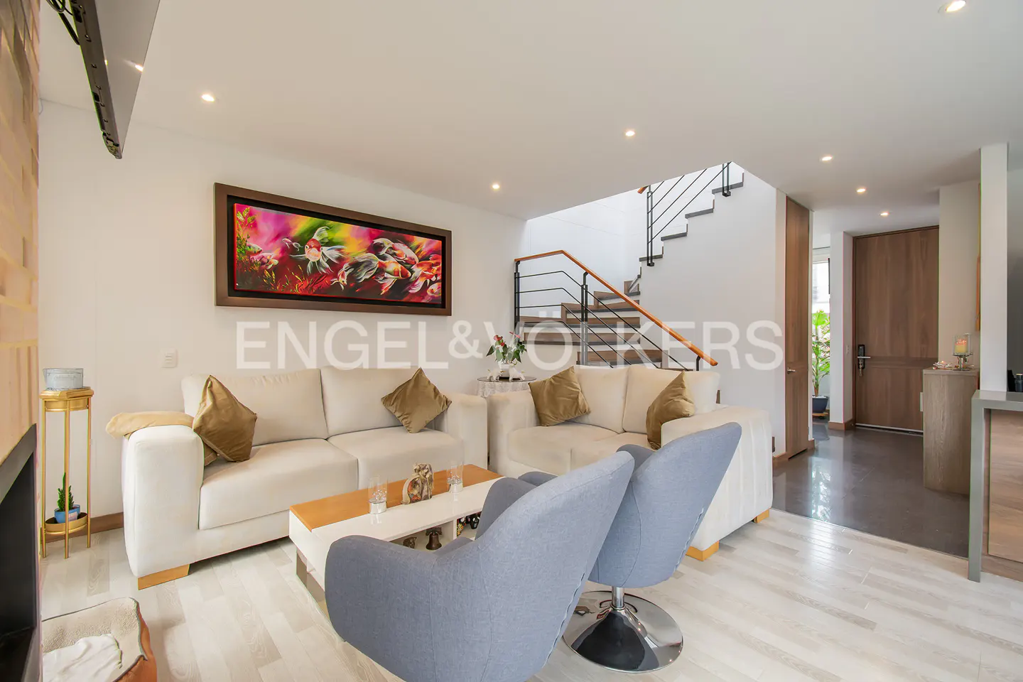 Bright living room with white sofas, blue chairs, and a wooden staircase. A colorful painting hangs above the sofa.