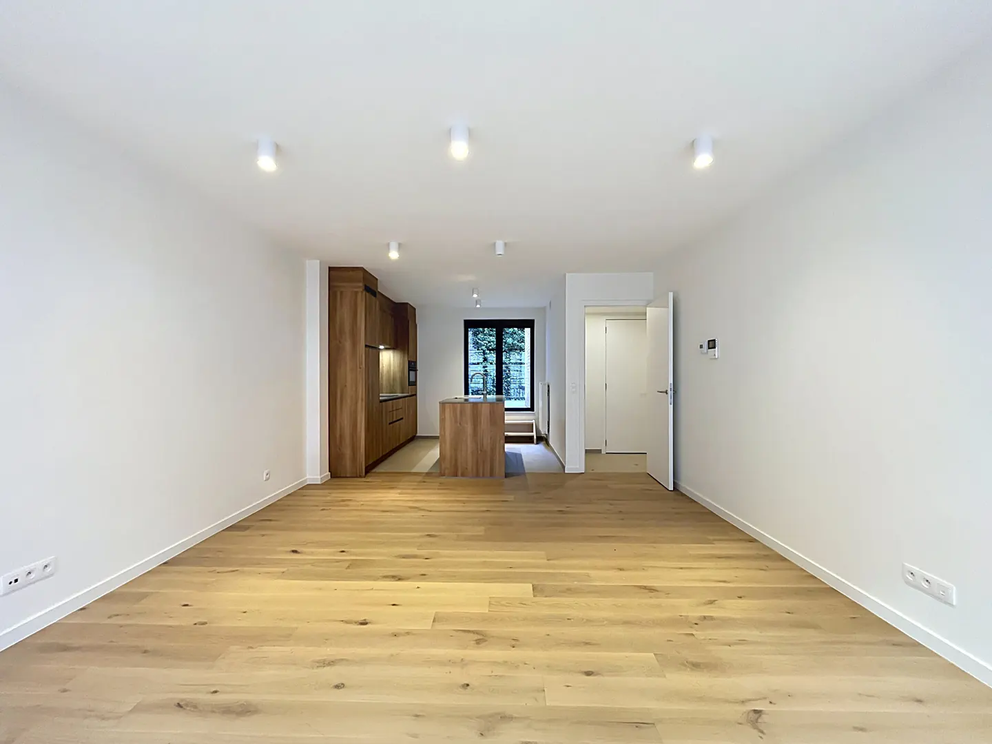 Bright, open-plan apartment with light wood floors, white walls, and recessed lighting. A wood kitchen and island are in the background.