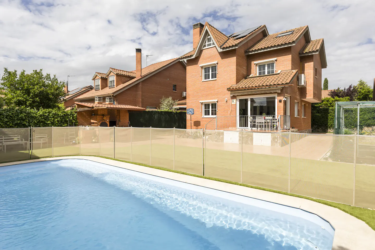 A backyard view of a two-story red brick house with a blue swimming pool and a beige safety fence.