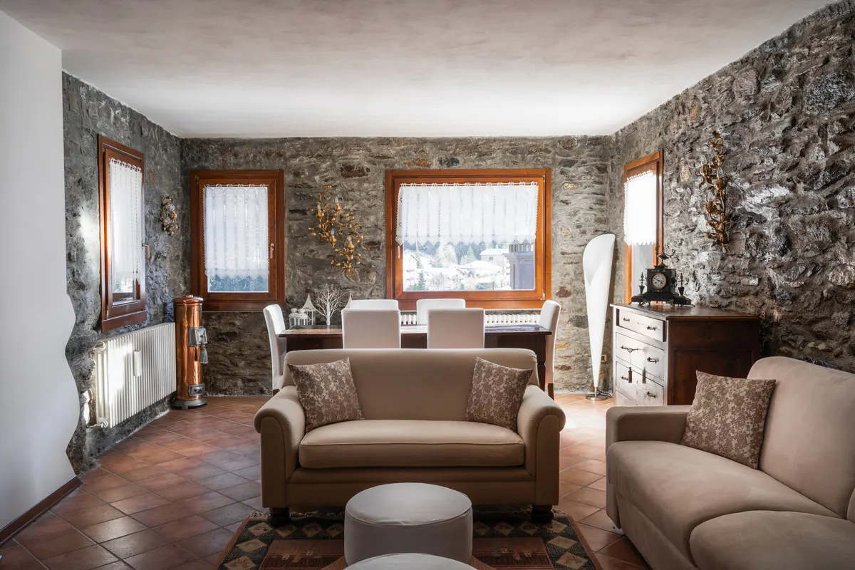 Living room with stone walls, terracotta tile floor, and beige sofas. Windows with white curtains overlook a snowy landscape.