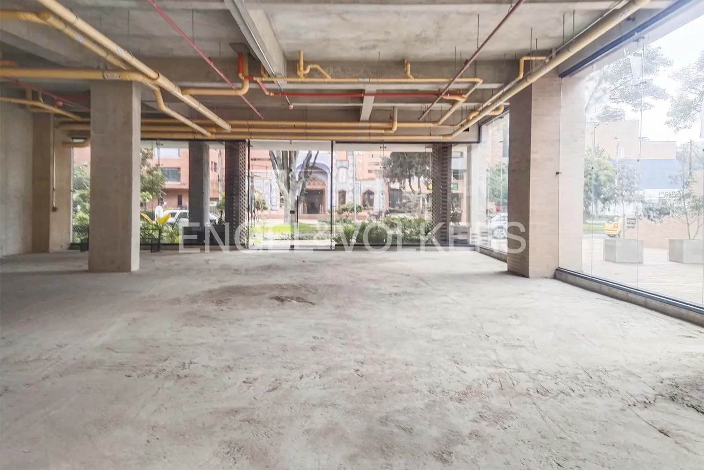 Empty commercial space with concrete floors, pillars, and exposed pipes. Large windows offer a view of a building and greenery.