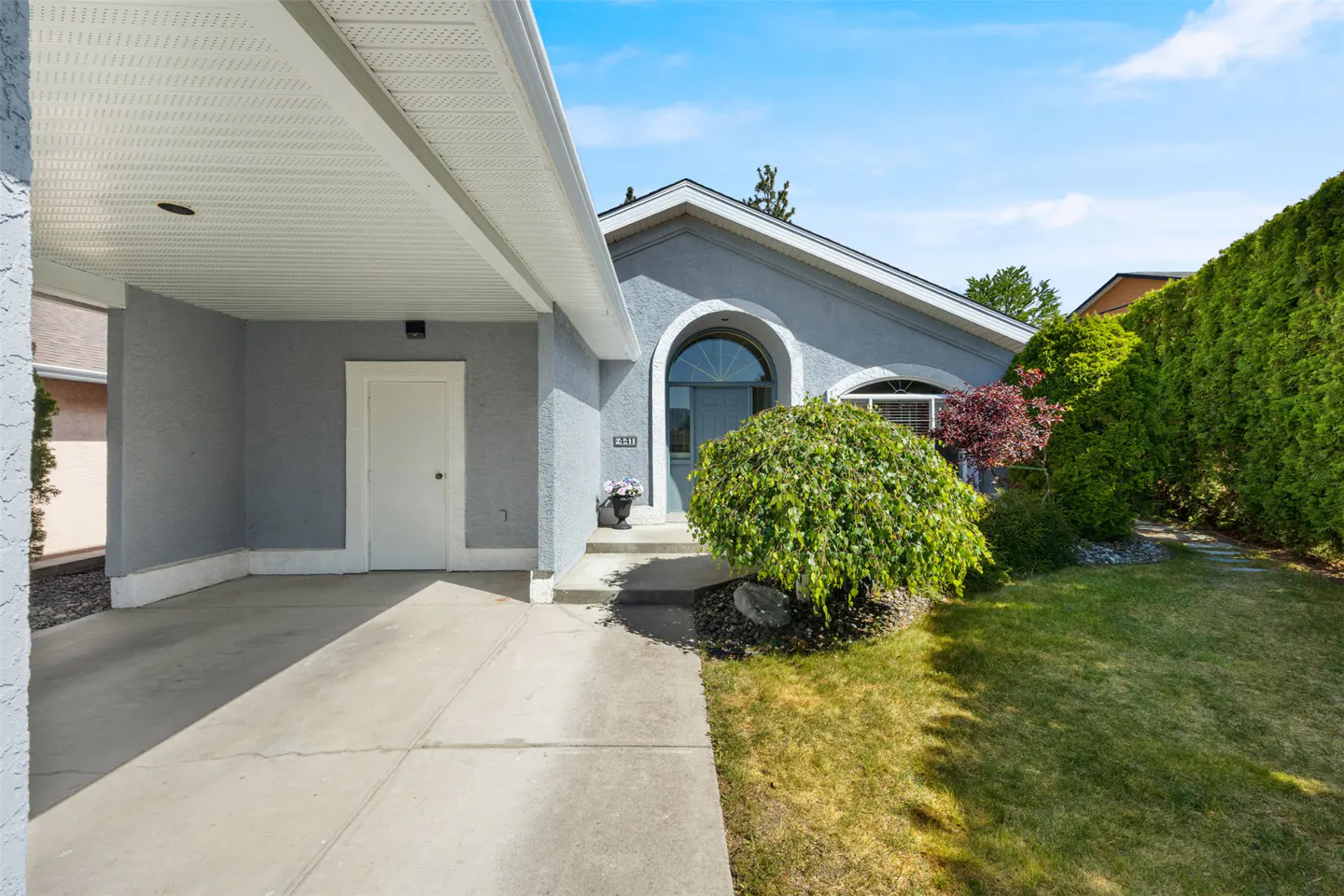 Exterior of a gray stucco house with a covered carport, arched entryway, and green lawn.