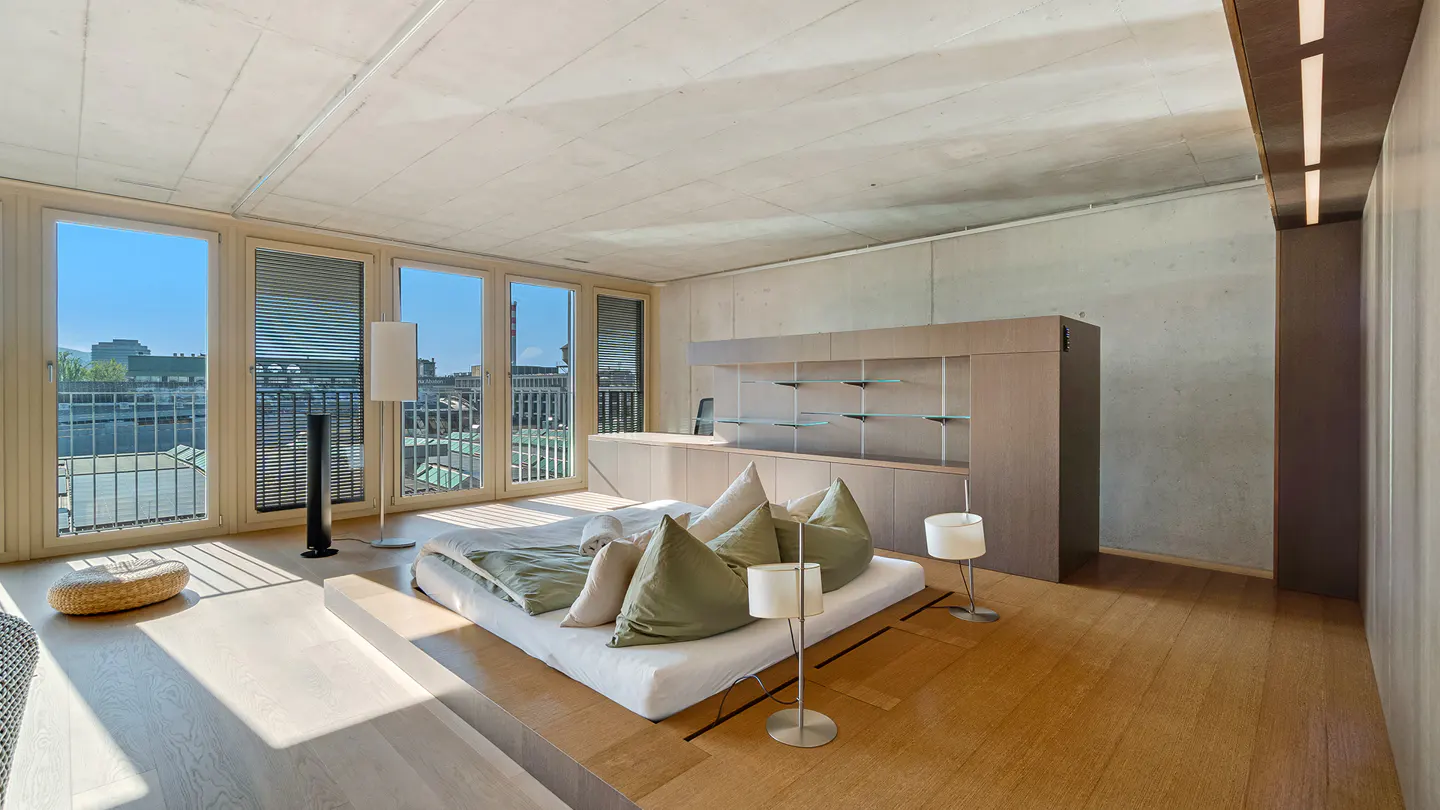 Bright bedroom with a low platform bed, green pillows, and floor lamps. Large windows offer a city view. Minimalist design with wood floors and a concrete ceiling.