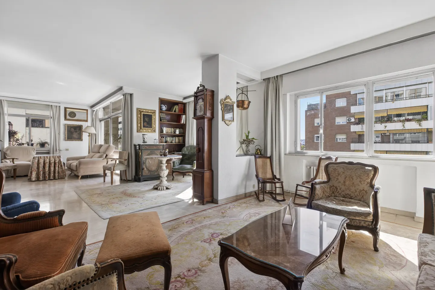 Bright, spacious living room with antique furniture, marble floors, and large windows overlooking a city building.