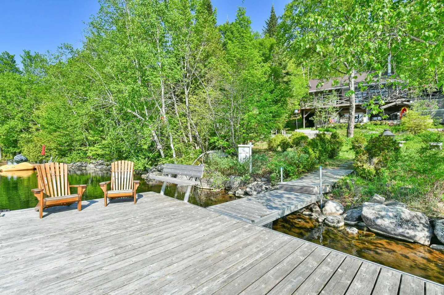 Wooden dock with two chairs overlooking a lake surrounded by green trees and a house in the background.