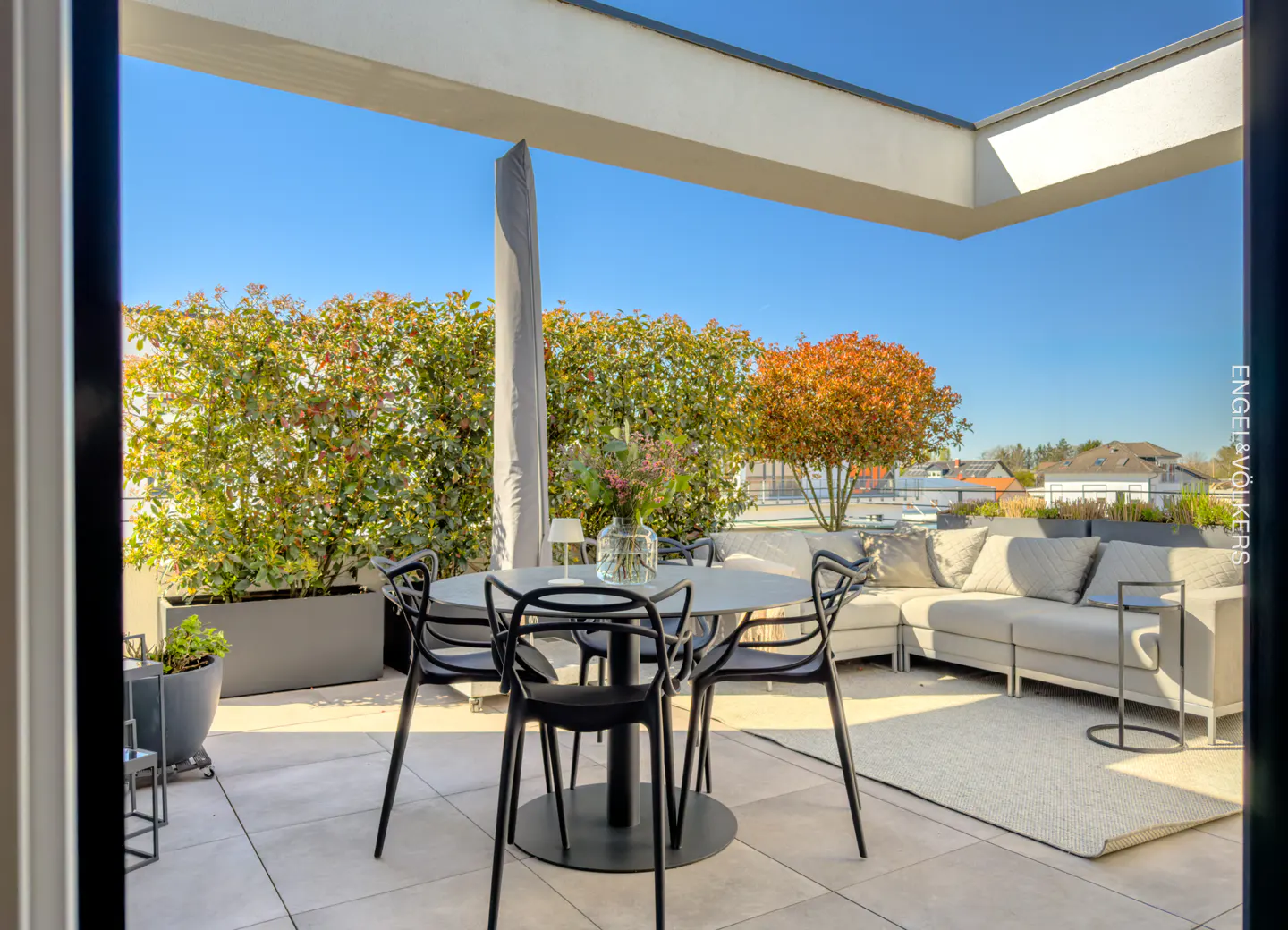Outdoor patio with a round table, black chairs, and a gray sofa with pillows. Green hedges and a blue sky are in the background.