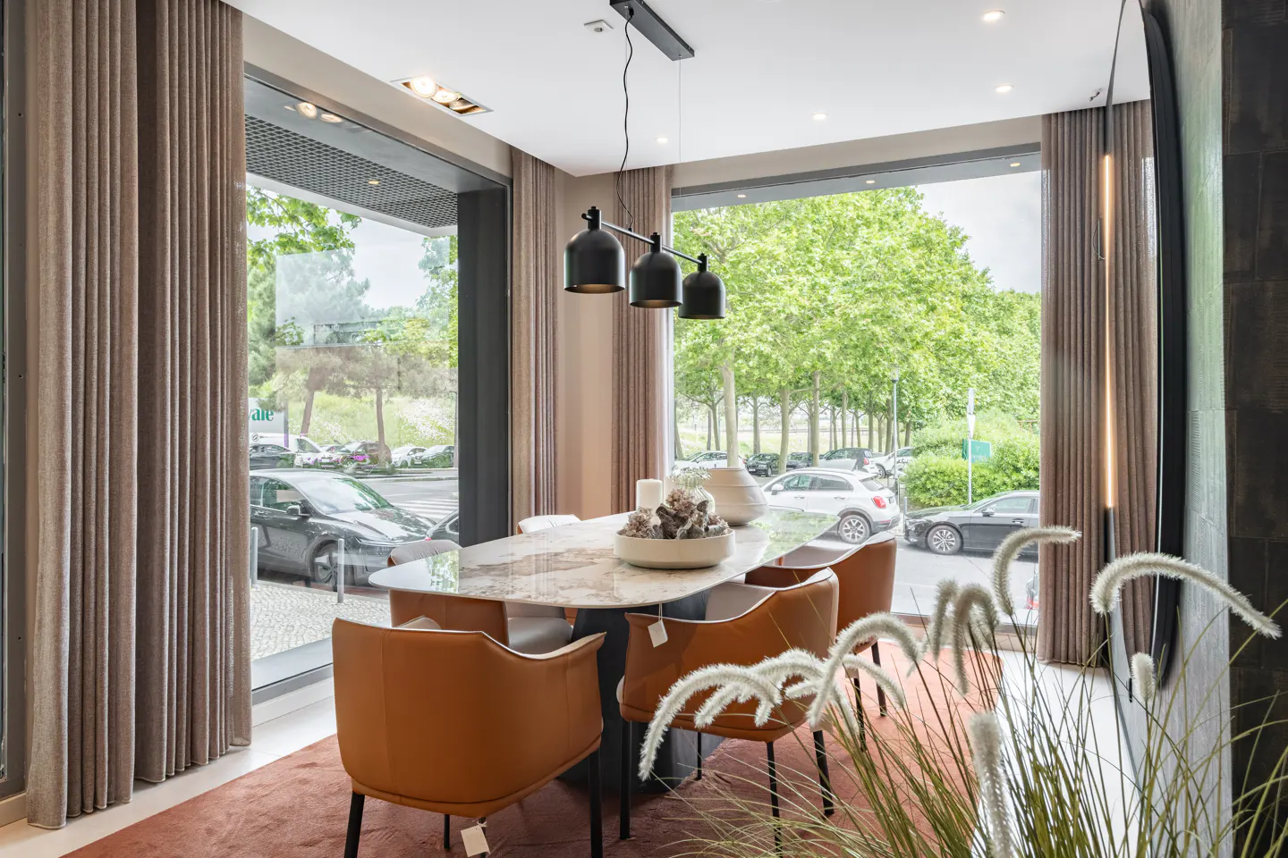 A modern dining room with a marble table, brown leather chairs, and a black pendant light. Large windows offer a view of a tree-lined street.