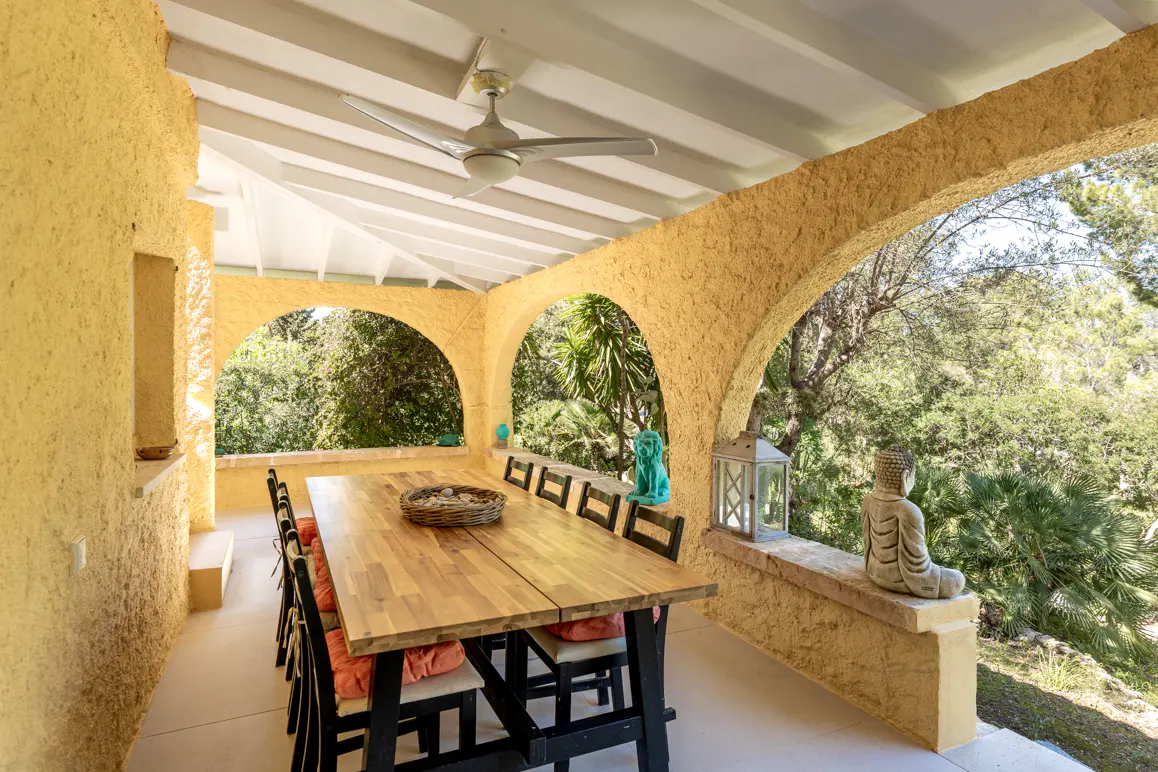 Covered patio with a long wooden table, black chairs with orange cushions, and arched openings overlooking lush greenery. A ceiling fan is overhead.