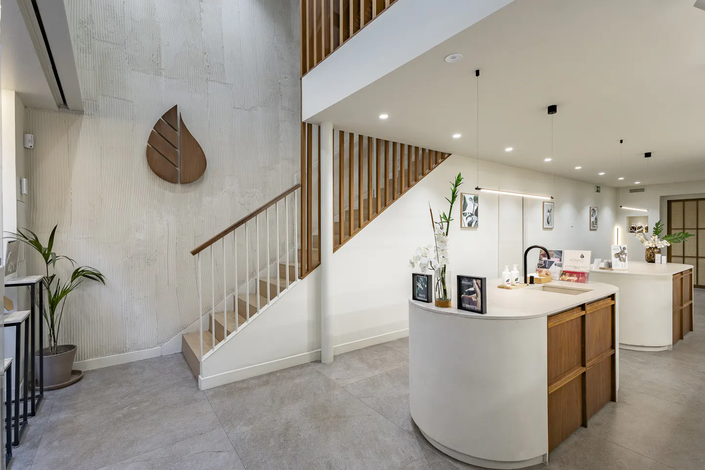 Modern office interior with white walls, wood accents, and gray tile flooring. A staircase leads to a second level. Reception desks are curved.