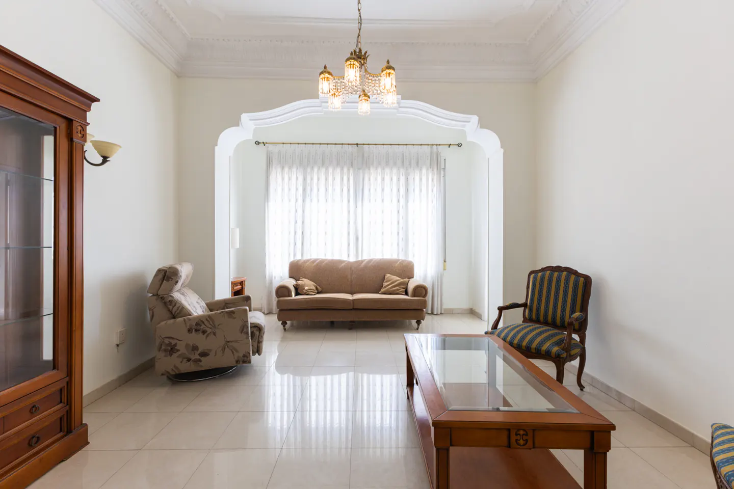 Bright living room with beige tile floor, white walls, and a tan sofa. A glass-topped coffee table sits in front of the sofa. A wooden cabinet is on the left.