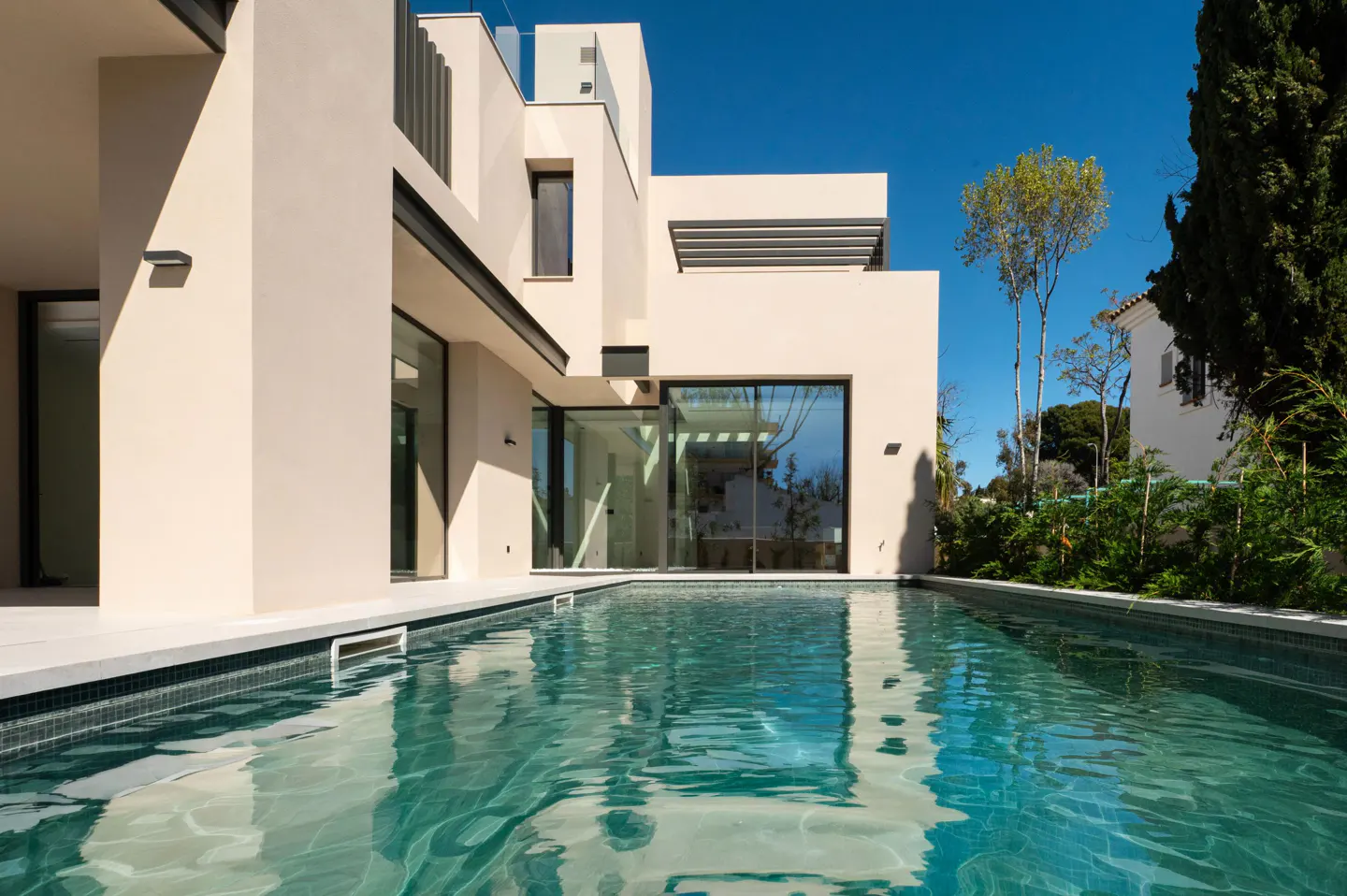 Modern beige house with a pool. Large glass doors reflect the sky. Trees and blue sky in the background.