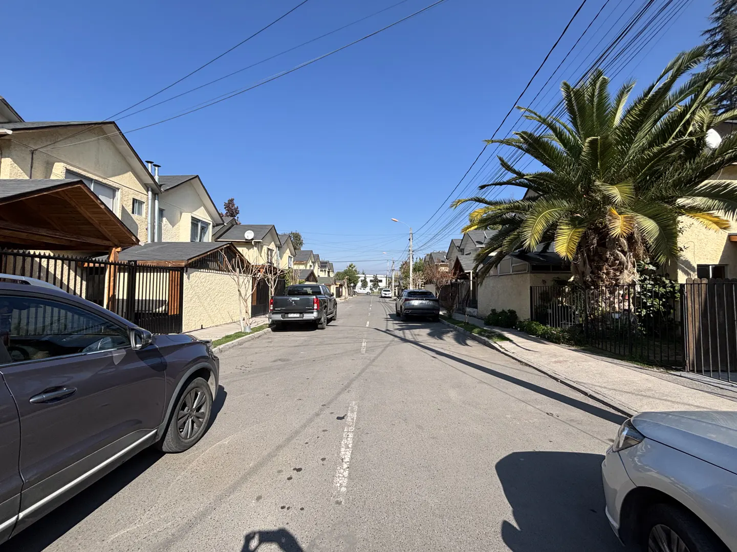Residential street view with parked cars, stucco houses, palm tree, and clear blue sky.