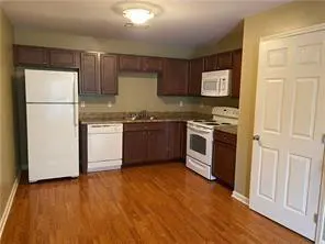 A kitchen with brown cabinets, white appliances, and wood floors. A white door is on the right.