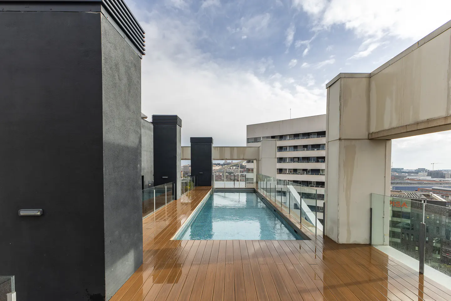Rooftop lap pool with wooden decking, glass railings, and modern black pillars against a city skyline.