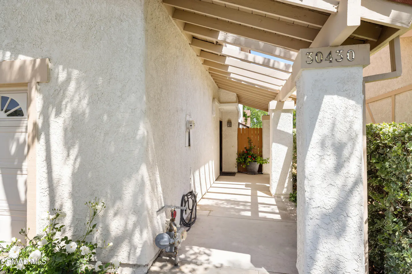 Exterior view of a house entrance with address "30430" on a pillar. A beige pergola casts shadows on the white stucco walls and concrete walkway. Green bushes and flowers add color.