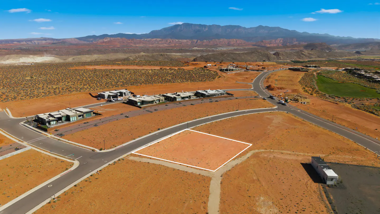 Aerial view of a vacant lot outlined in white, surrounded by desert landscape, modern homes, and distant mountains.