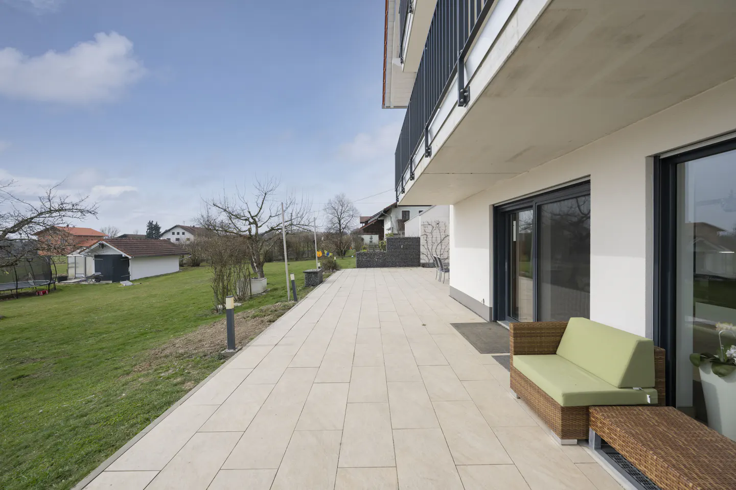 A patio with a green sofa and a wicker table, next to a white building with black framed windows, overlooking a green lawn.