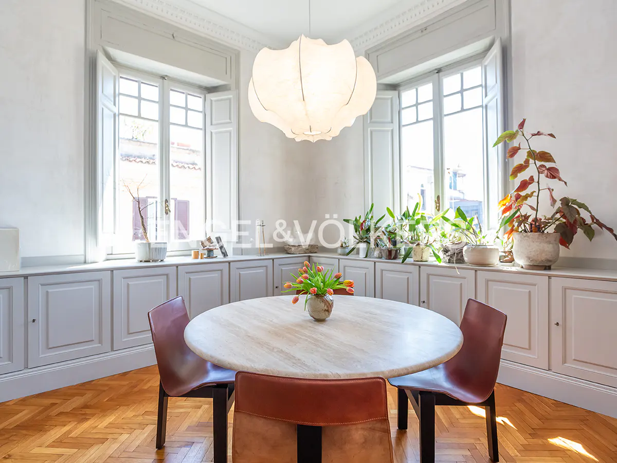 Bright dining room with a round table, four chairs, and a large white pendant light. Windows with plants line the back wall.