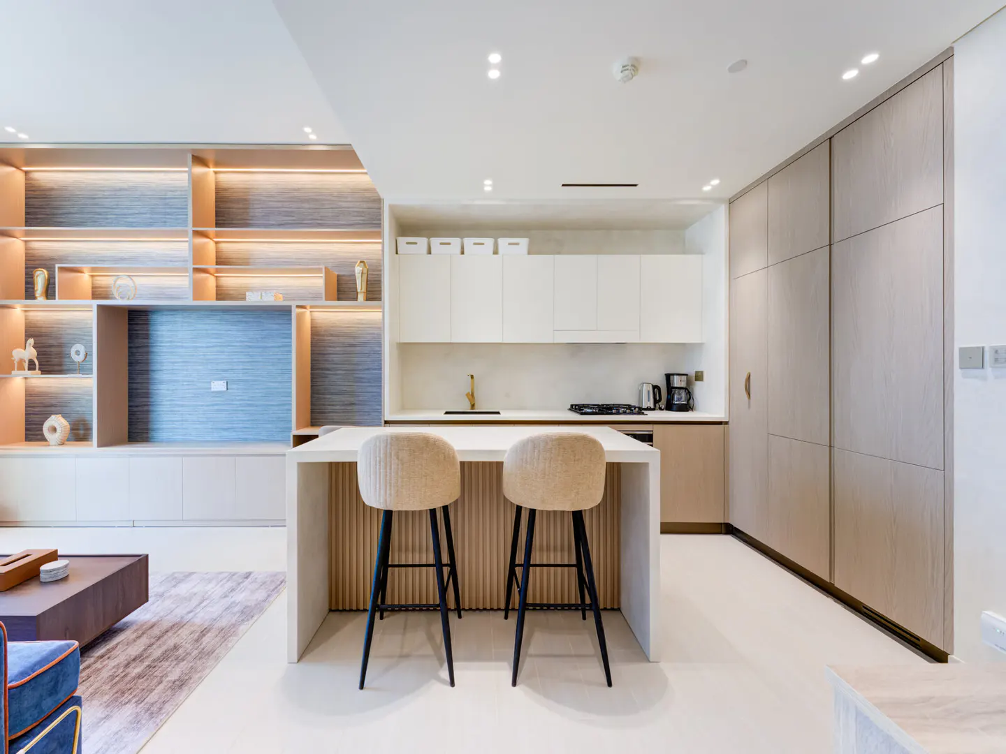 Modern kitchen with island and two stools. White cabinets, wood accents, and built-in shelving with decorative items. Bright, open space.
