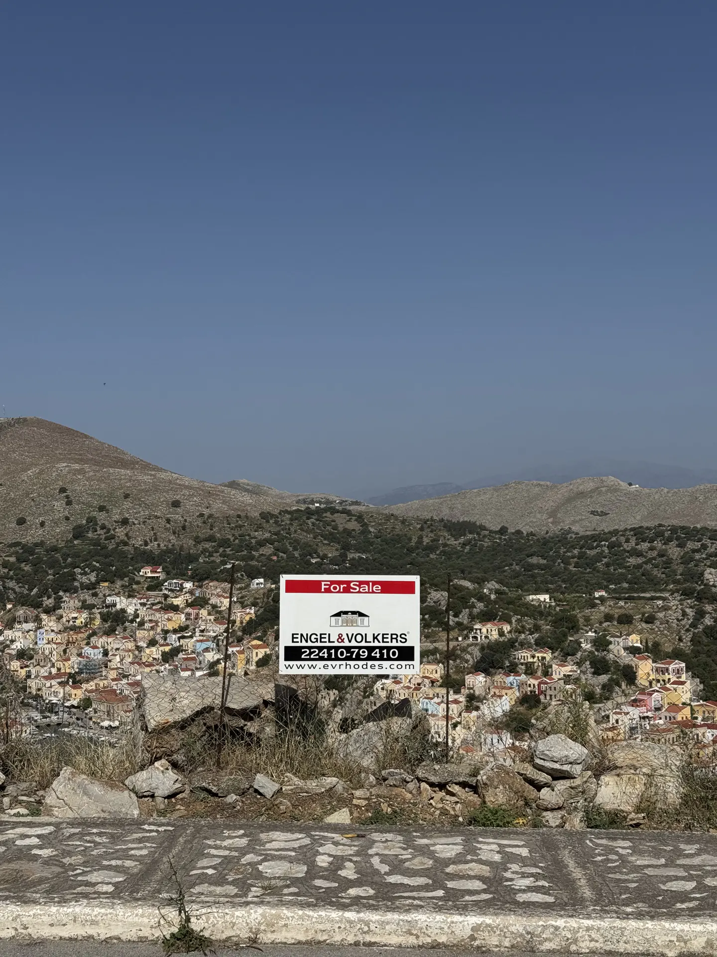 Engel & Völkers for sale sign overlooking a hillside village with colorful houses under a clear blue sky.