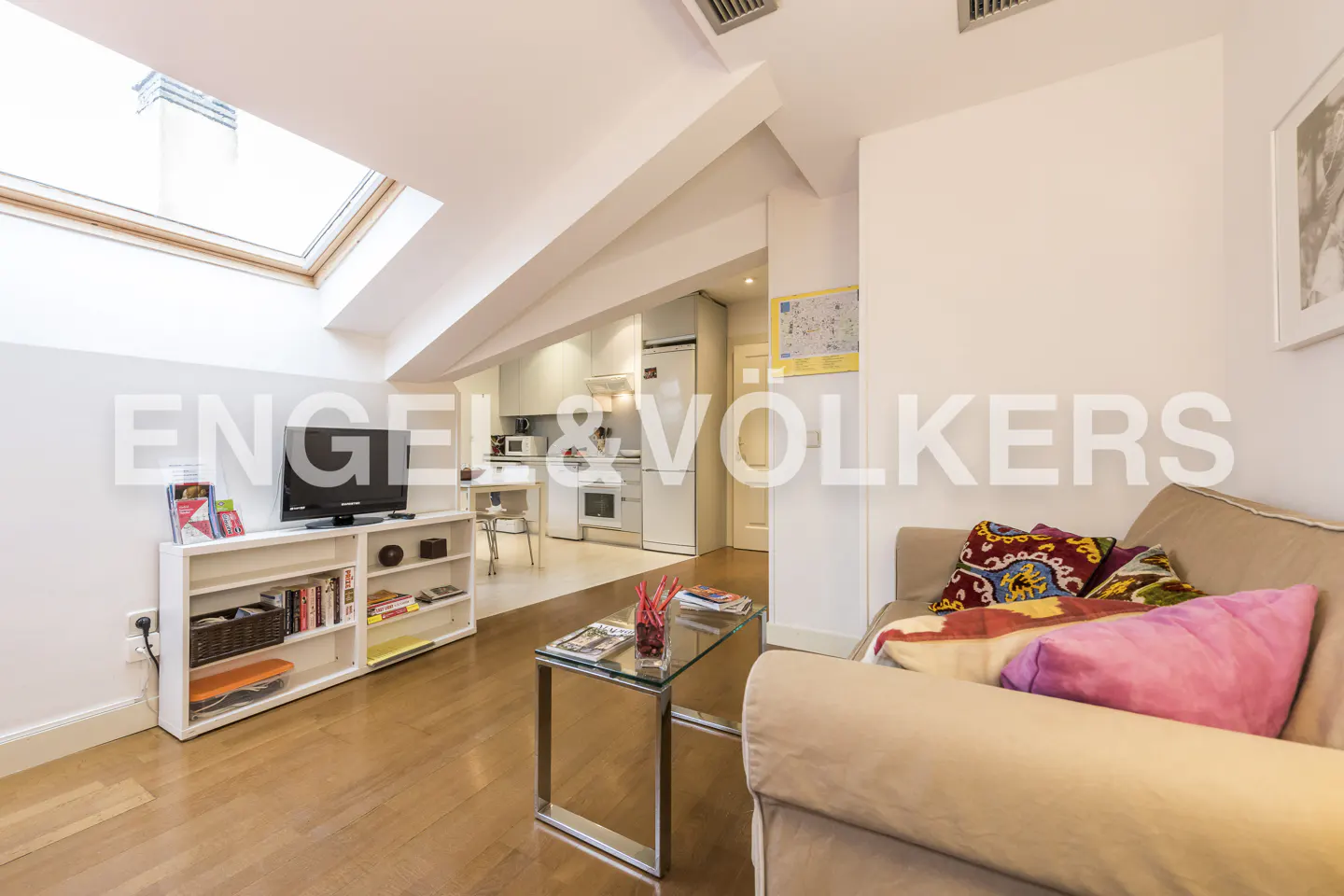 Living room with wood floors, a beige sofa with colorful pillows, a glass coffee table, and a white bookshelf with a TV. A skylight is visible.