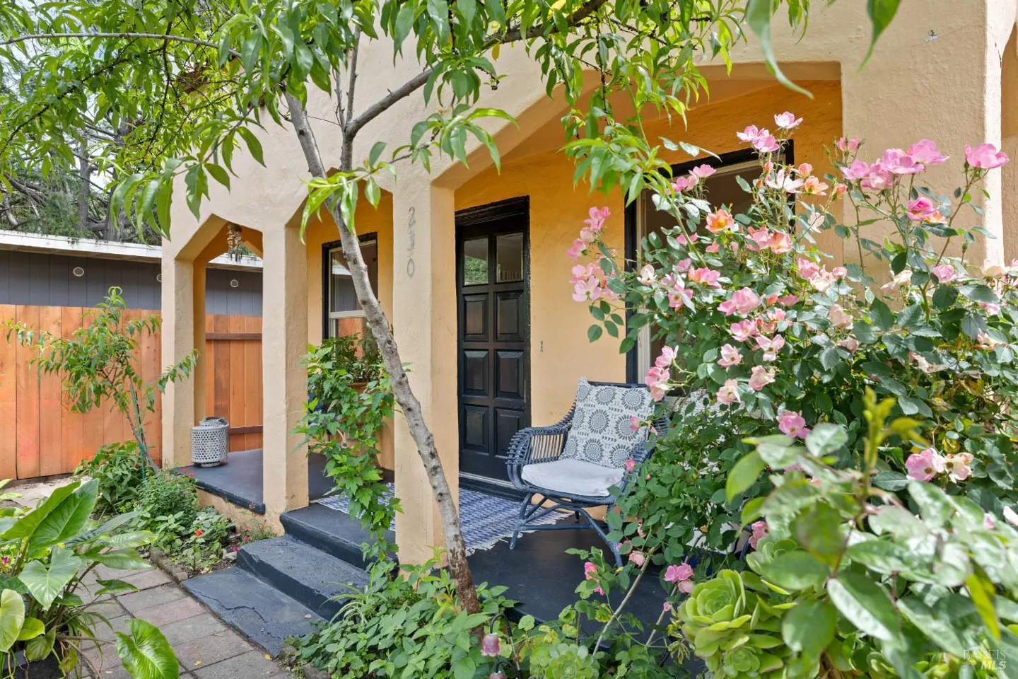 Charming home exterior with a black door, tan stucco, and pink roses. A wicker chair sits on the porch, inviting relaxation. Lush greenery surrounds the entrance.