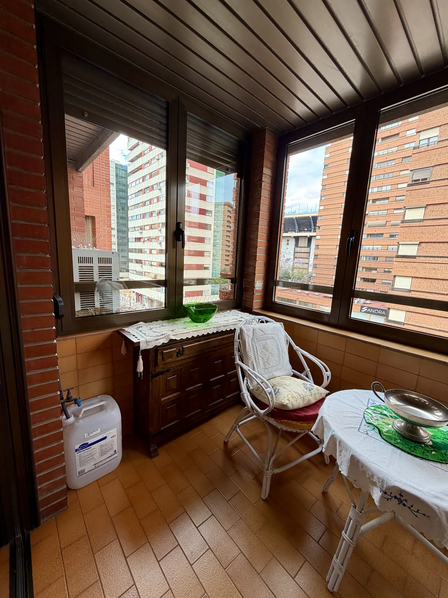 Indoor balcony with brick walls, brown tile floor, and large windows showing city buildings. A wooden chest, white wicker chair, and table furnish the space.