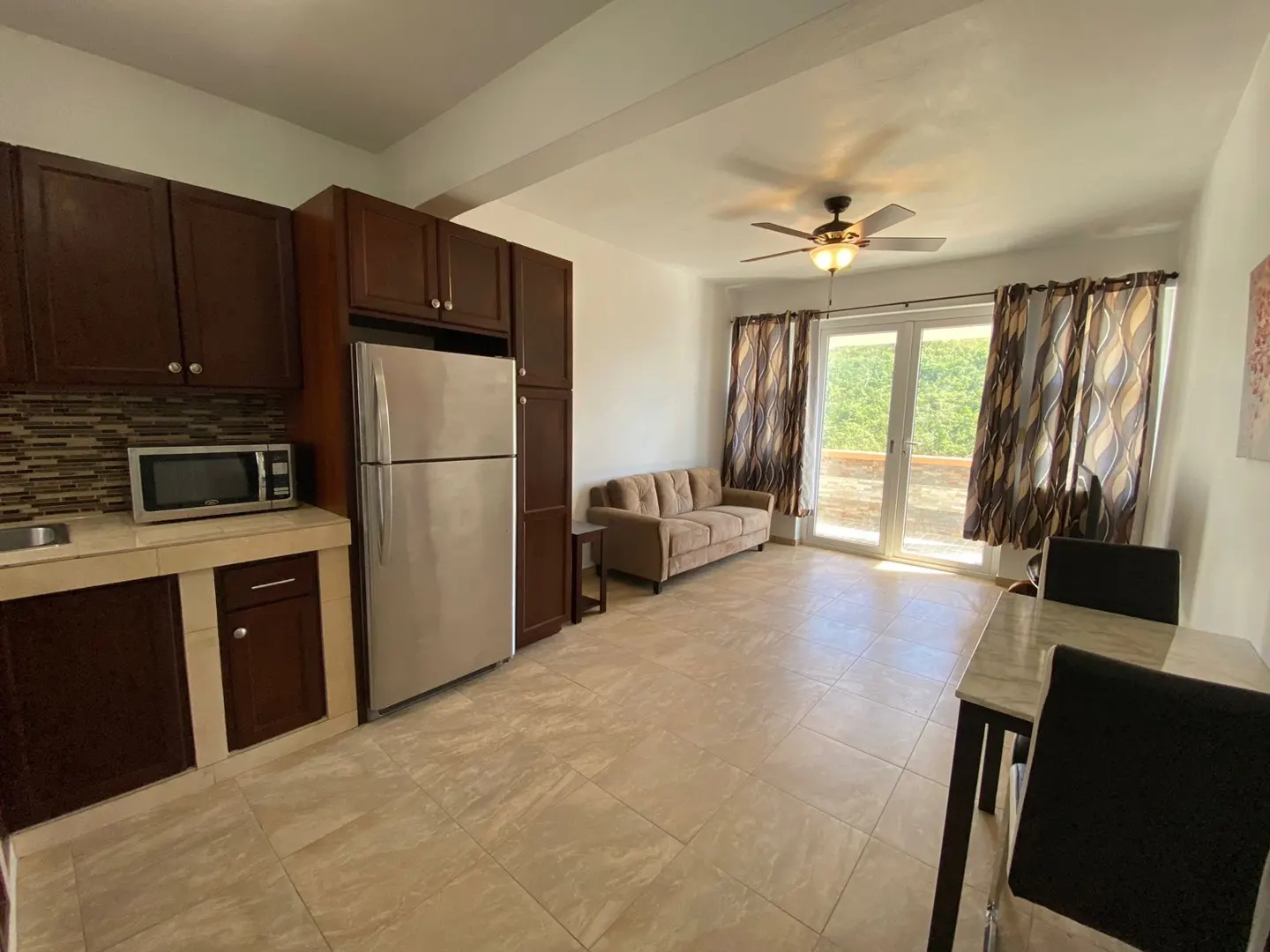 Open-concept living space with brown cabinets, stainless steel appliances, tan sofa, and dining table. Sliding glass doors lead to a balcony.