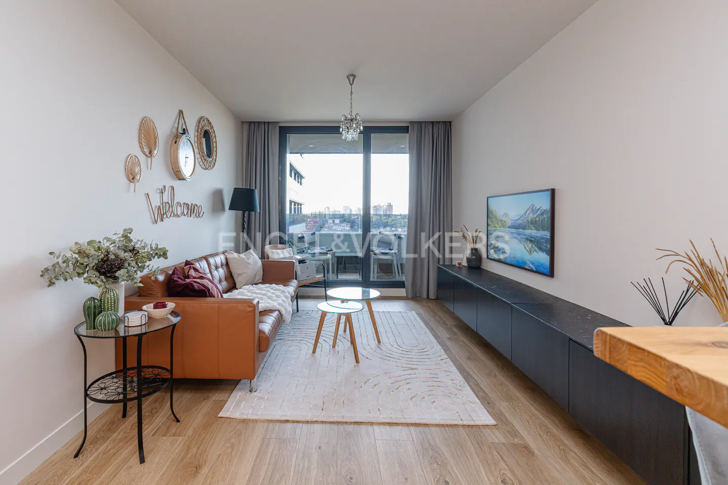 Modern living room with a tan leather sofa, two small round coffee tables, a black media console, and a balcony view.
