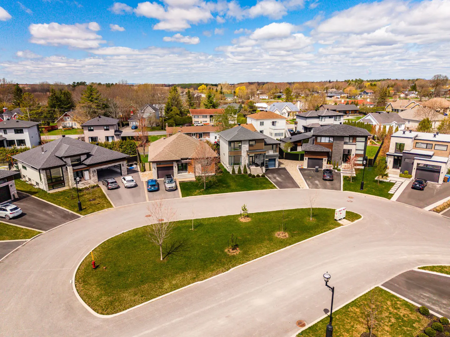 Aerial view of a suburban neighborhood with modern houses, green lawns, and a winding street under a blue sky with white clouds.