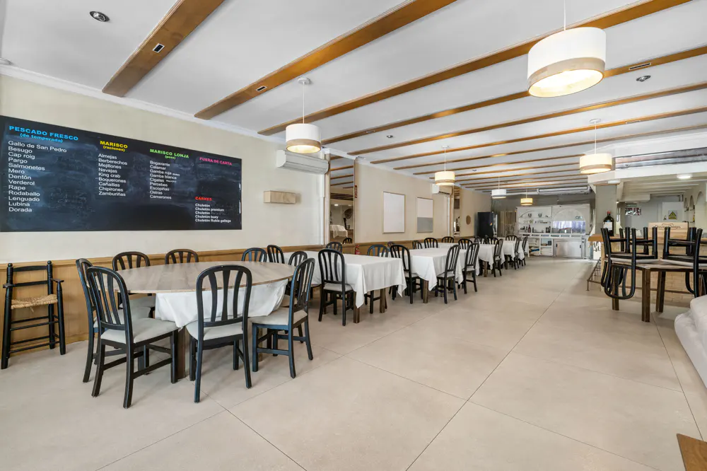 Restaurant interior with tables and chairs, a chalkboard menu, and overhead lighting. The floor is tiled and the ceiling has wooden beams.