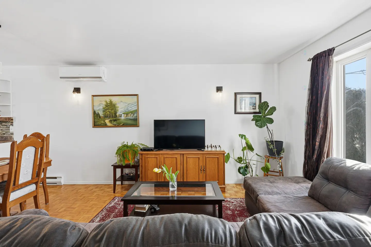 Living room with brown sofa, wood floors, and white walls. A TV sits on a wooden cabinet, with plants and artwork adding color.