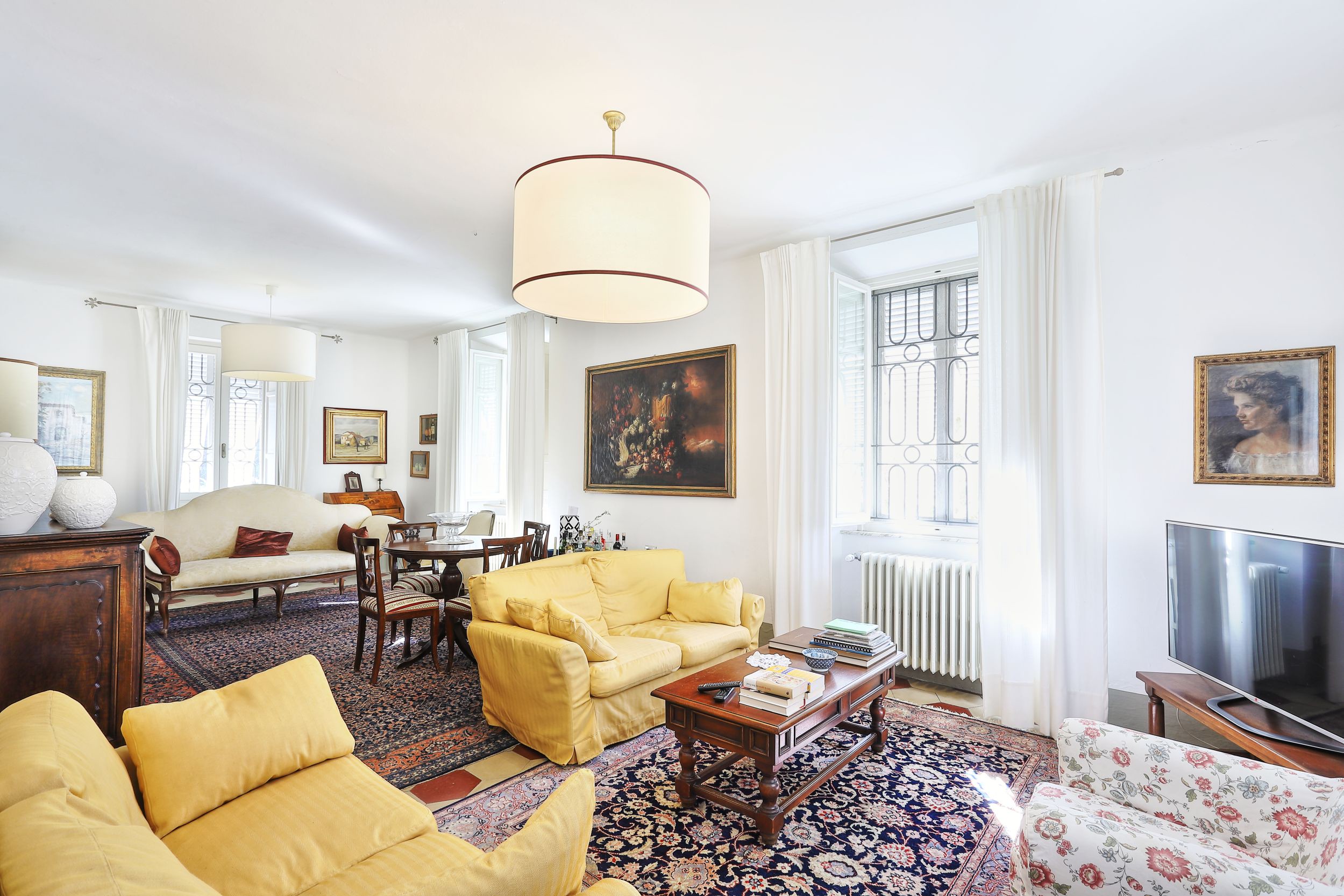 Bright living room with yellow sofas, a round wooden table, and patterned rugs. White walls and curtains add to the airy feel. [AI-generated text]