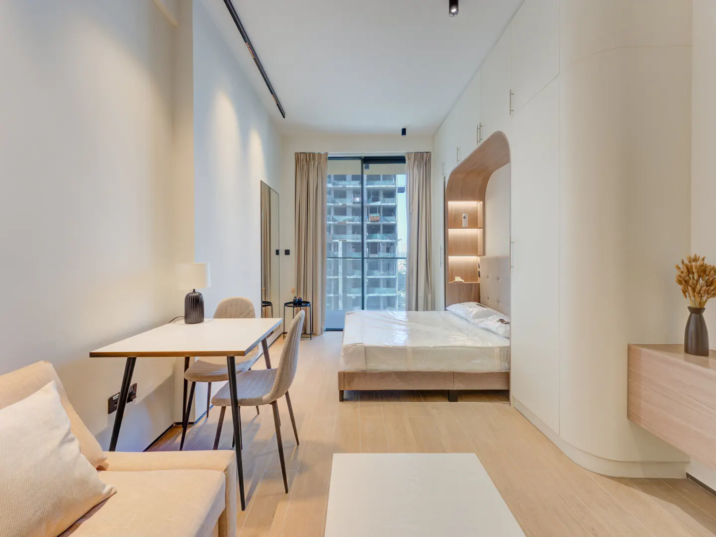 Bright studio apartment with a dining table, sofa, and bed nook with built-in shelving and a view of a city building.