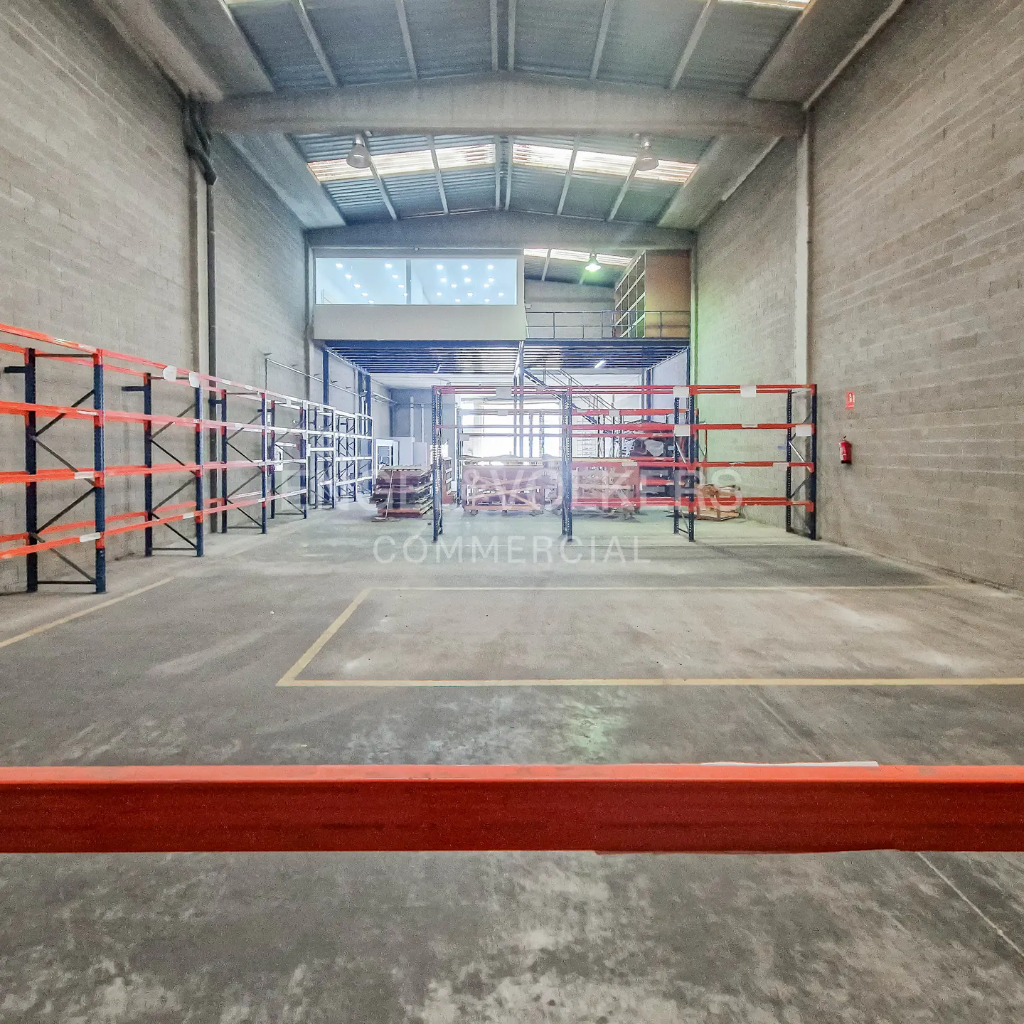 A large, empty warehouse with concrete floors and walls, metal shelving, and a mezzanine level.
