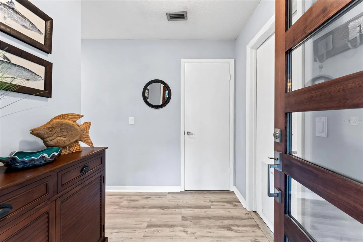 Entryway with wood-look floors, light blue walls, and a dark wood cabinet with fish decor. A modern wood and glass door is ajar.
