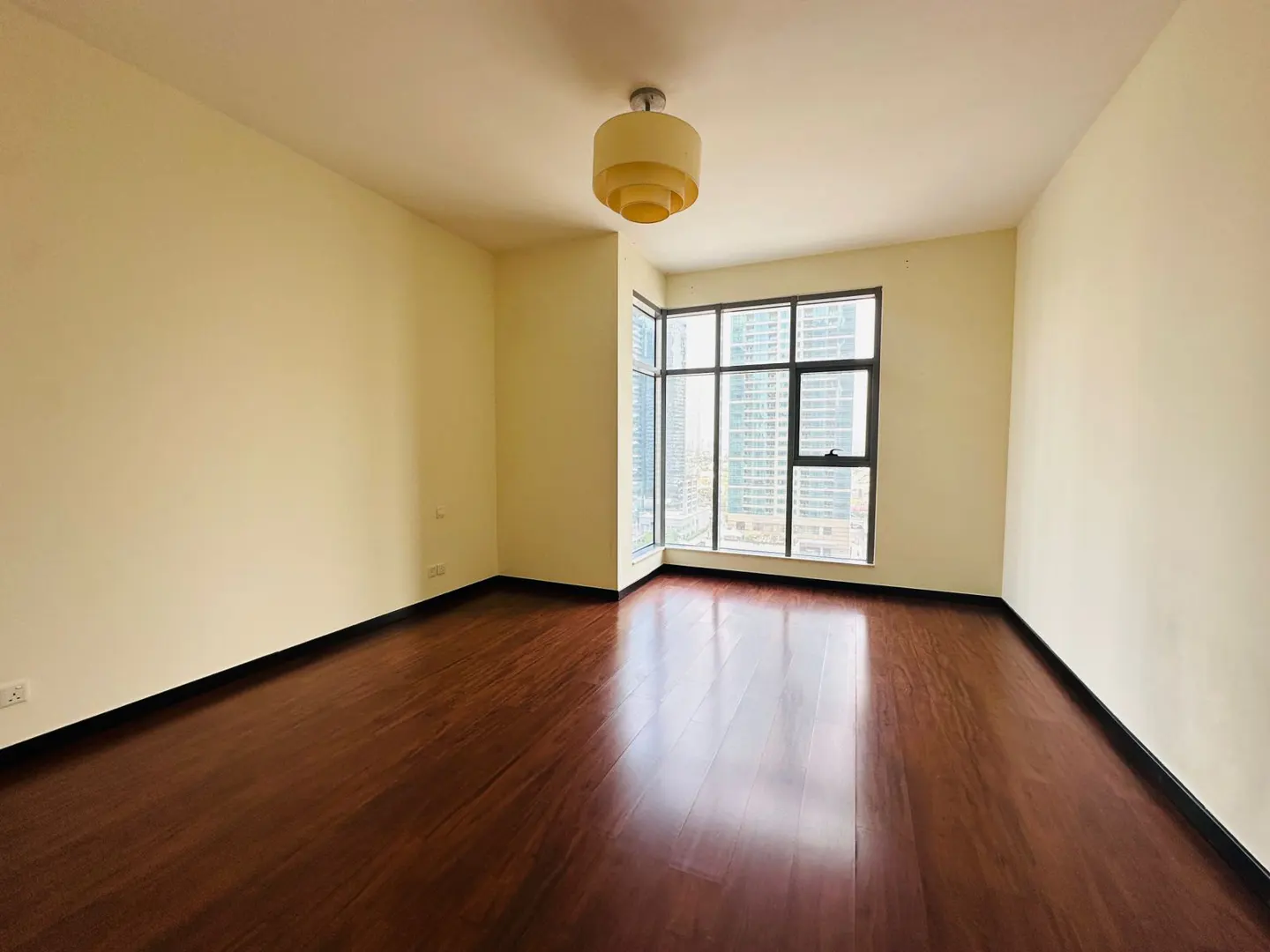 Empty room with dark wood floors, light yellow walls, and a large window with a city view. A tiered light fixture hangs from the ceiling.