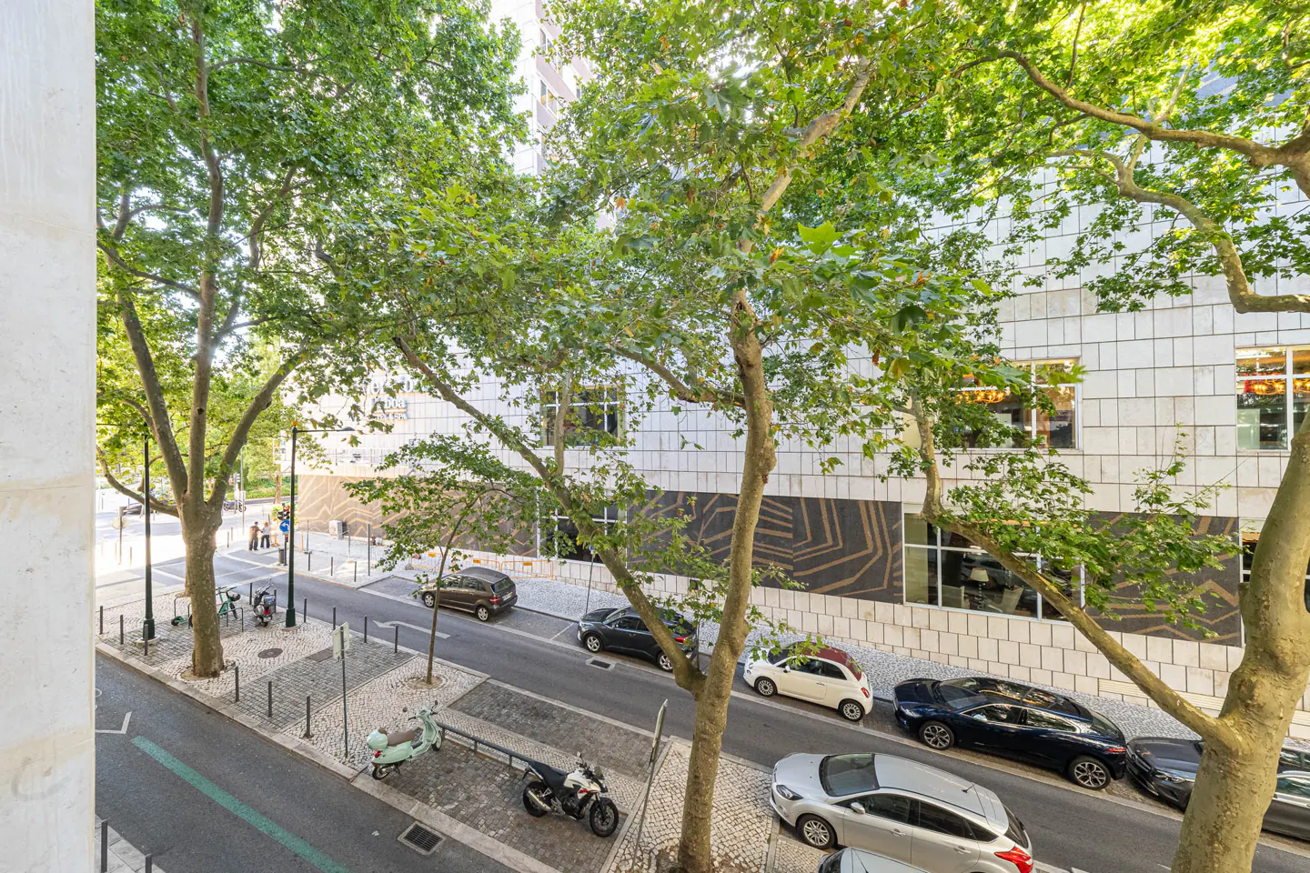 High-angle view of a city street with parked cars, a scooter, and a modern building facade framed by large green trees.