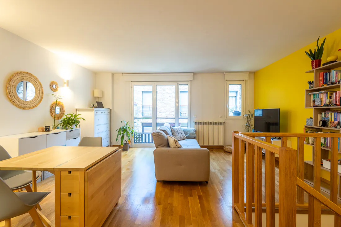 Bright living room with wood floors, a beige sofa, and a wooden dining table with gray chairs. A yellow accent wall features a bookshelf and TV.