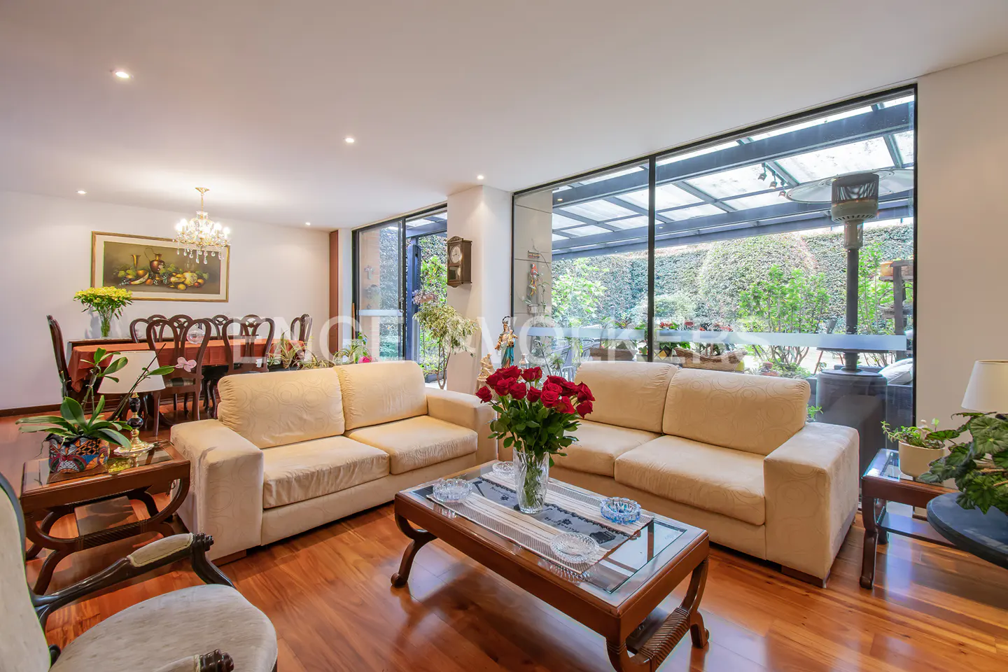 Living room with two beige sofas, a glass coffee table with red roses, and a dining table with chairs in the background. Large windows overlook a garden.