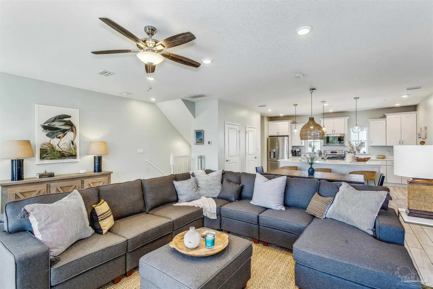 Open-concept living room with a gray sectional sofa, ottoman, and a ceiling fan. The kitchen has white cabinets and stainless steel appliances.