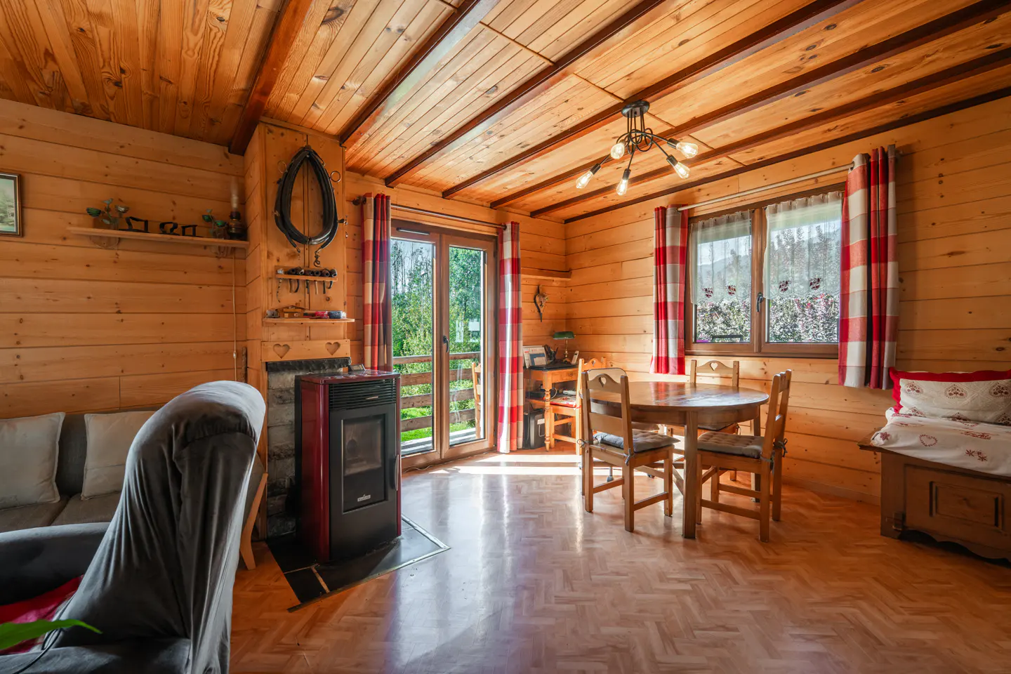 Cozy wood cabin interior with a round table, chairs, a red pellet stove, and a gray sofa. Red and white curtains adorn the windows.