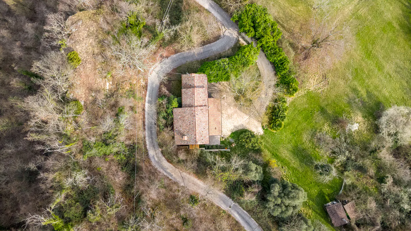 Aerial view of a house with a red tile roof, surrounded by trees and a winding road. A green field is visible on the right.