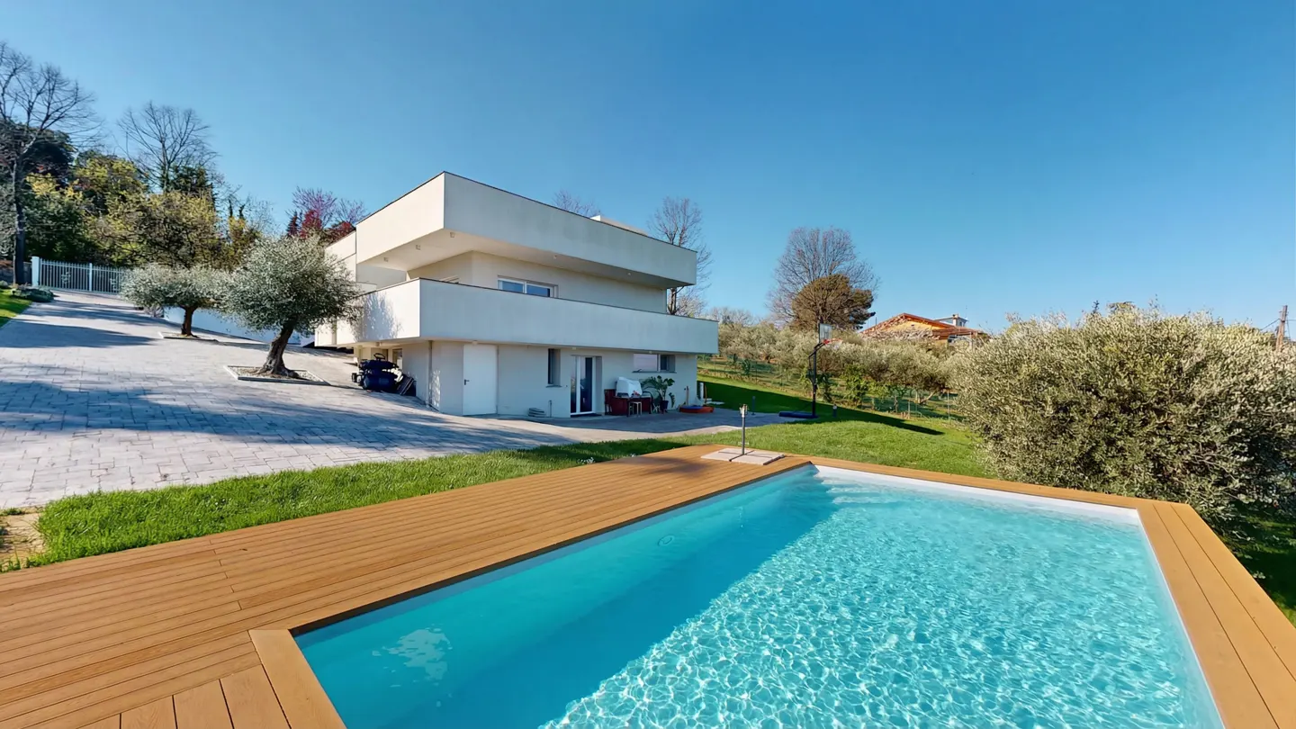 Modern white house with a pool. The pool has a wooden deck. Green grass and trees surround the property. Blue sky.